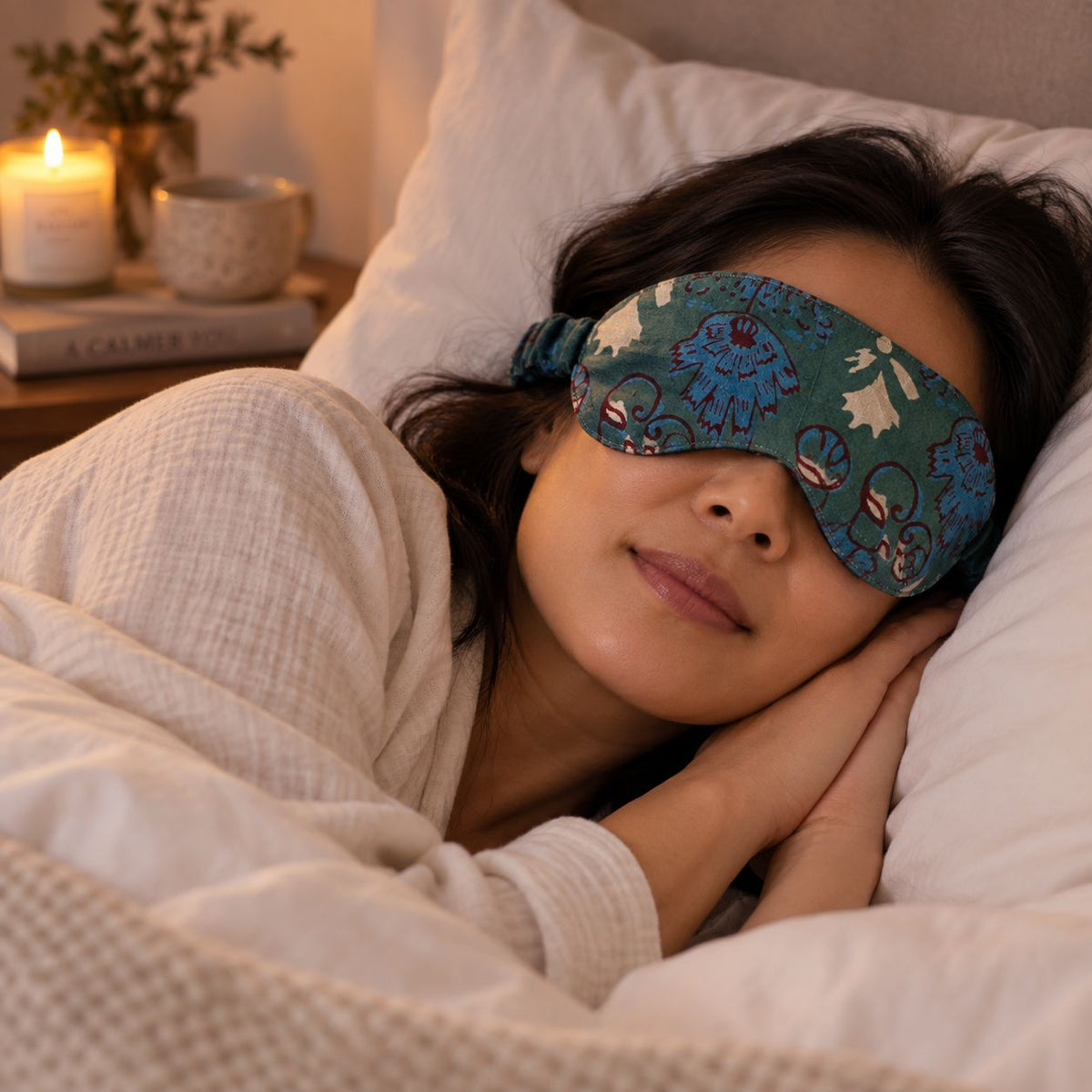Woman wearing the cotton sleep mask in bed with candle and book on bedside table.