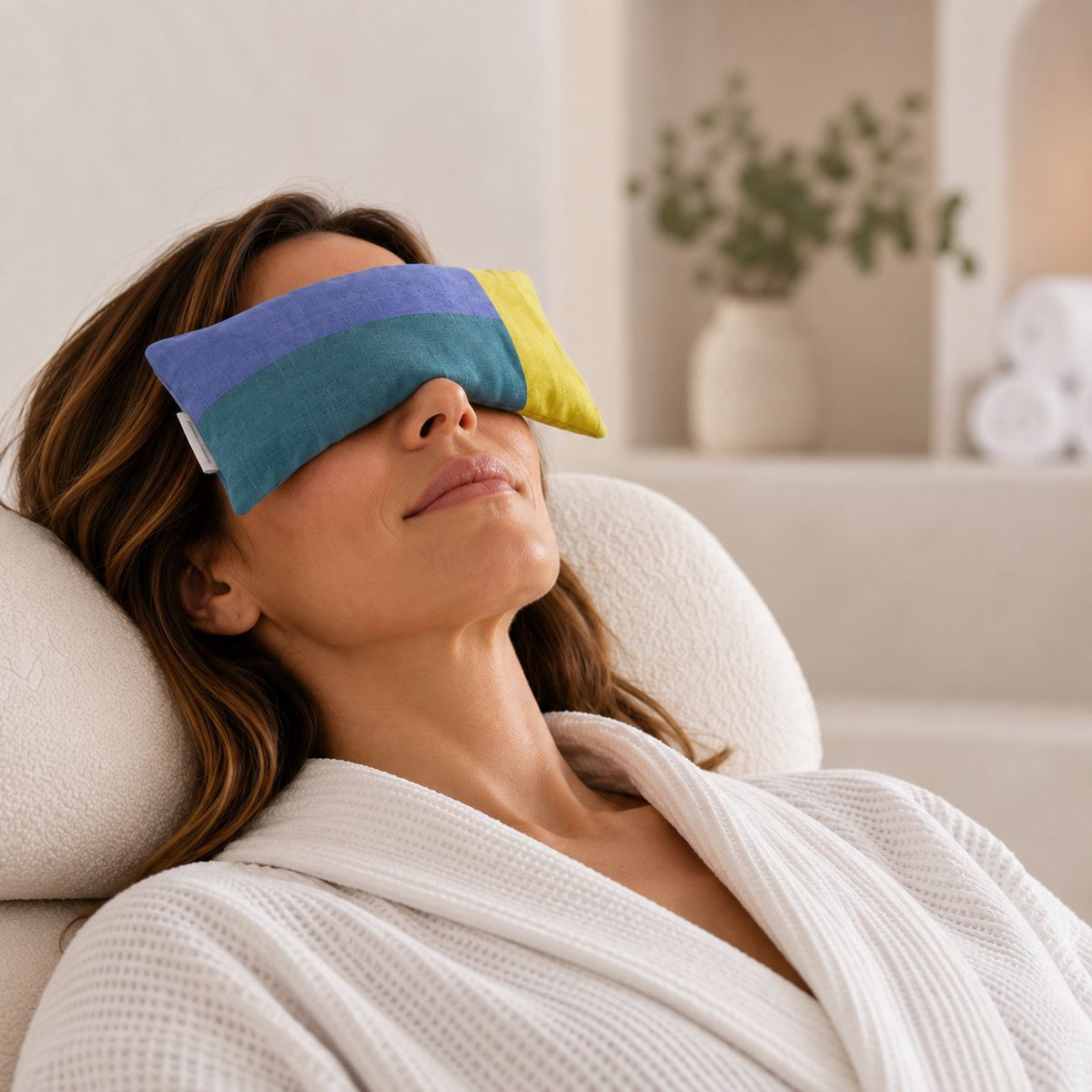 A woman reclining in a spa-like setting with a weighted eye pillow over her eyes, wearing a white robe.