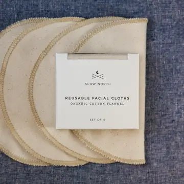 Four reusable cotton facial cloths in a cream color with a fringed edge, stacked on a blue textured fabric background with a white paper label in the center reading 'SLOW NORTH REUSABLE FACIAL CLOTHS ORGANIC COTTON PLANNEL SET OF 4'.