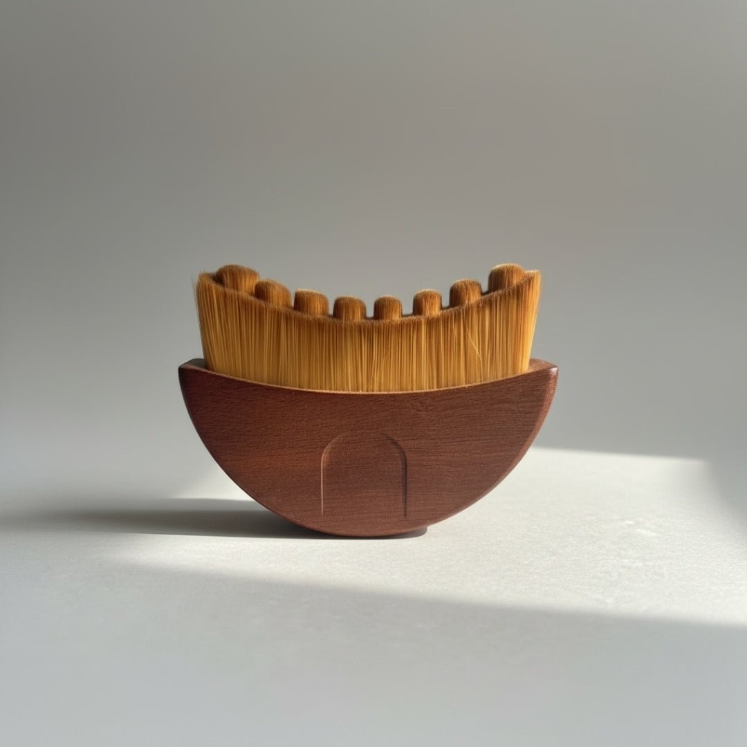 A gua sha brush with a wooden handle and soft, fine bristles in a crescent-shape arrangement on a light gray studio background with a soft shadow.