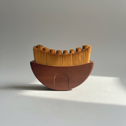 A gua sha brush with a wooden handle and soft, fine bristles in a crescent-shape arrangement on a light gray studio background with a soft shadow.