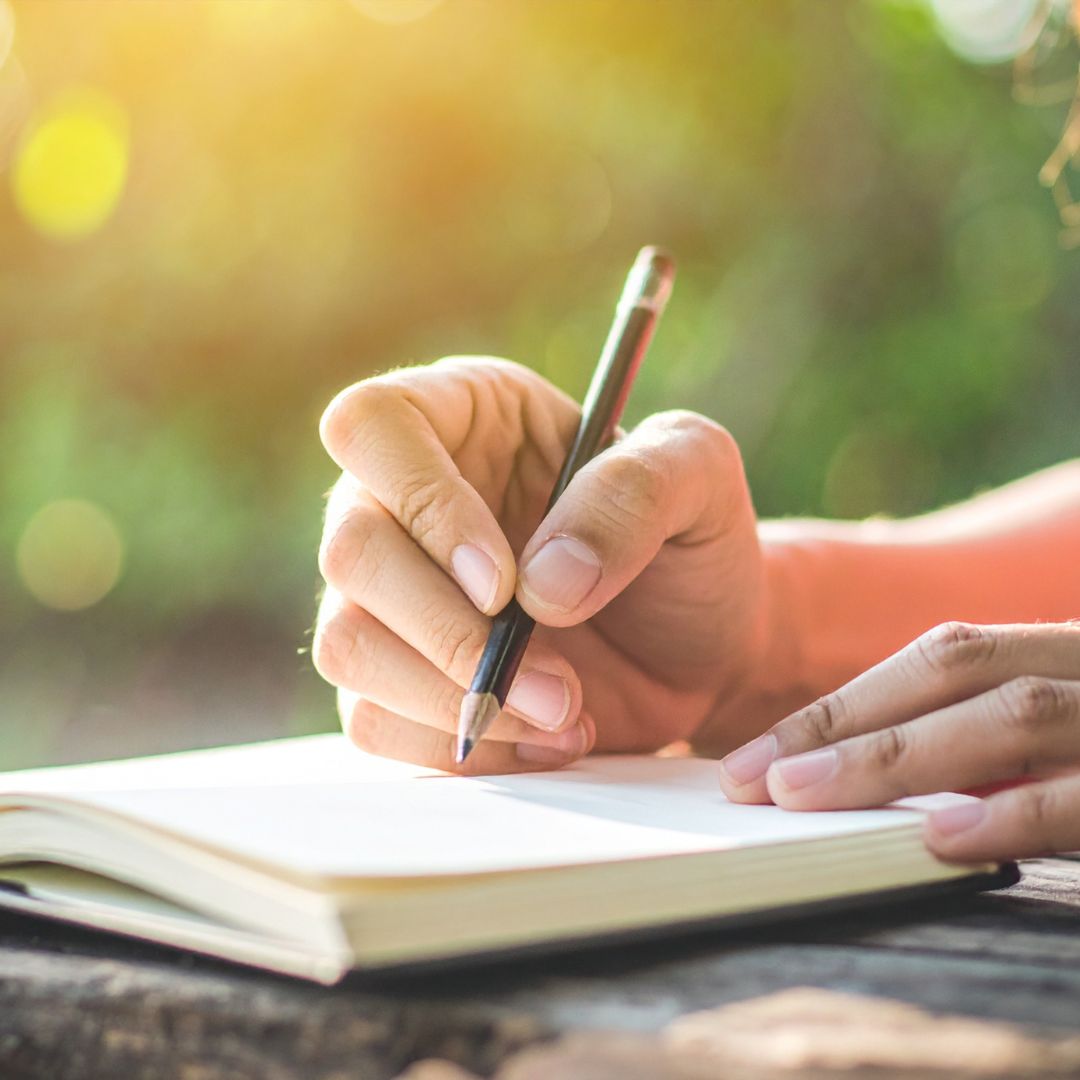 Hand writing in a notebook with a pencil, outdoors.