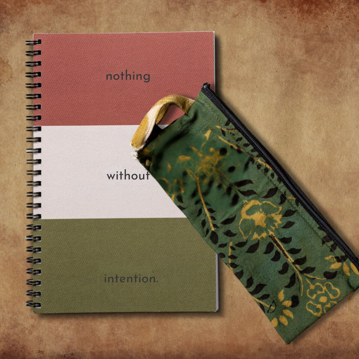 A spiral-bound notebook with a color-block cover and a matching fabric zip pouch with a green and gold floral pattern, arranged on a textured brown background.