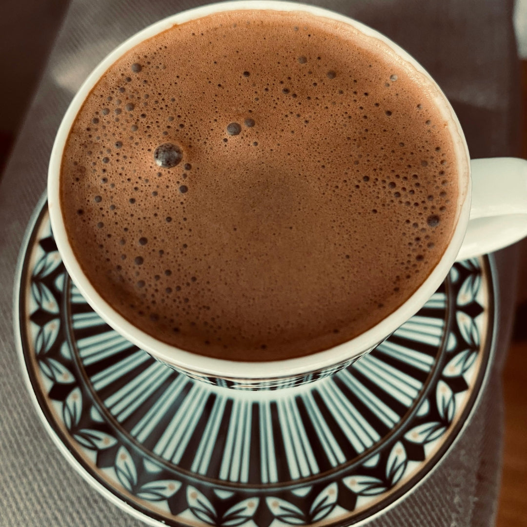 A top-down view of a white ceramic cup filled with frothy hot cacao drinking chocolate, resting on a matching saucer with a black, white, and blue geometric pattern.