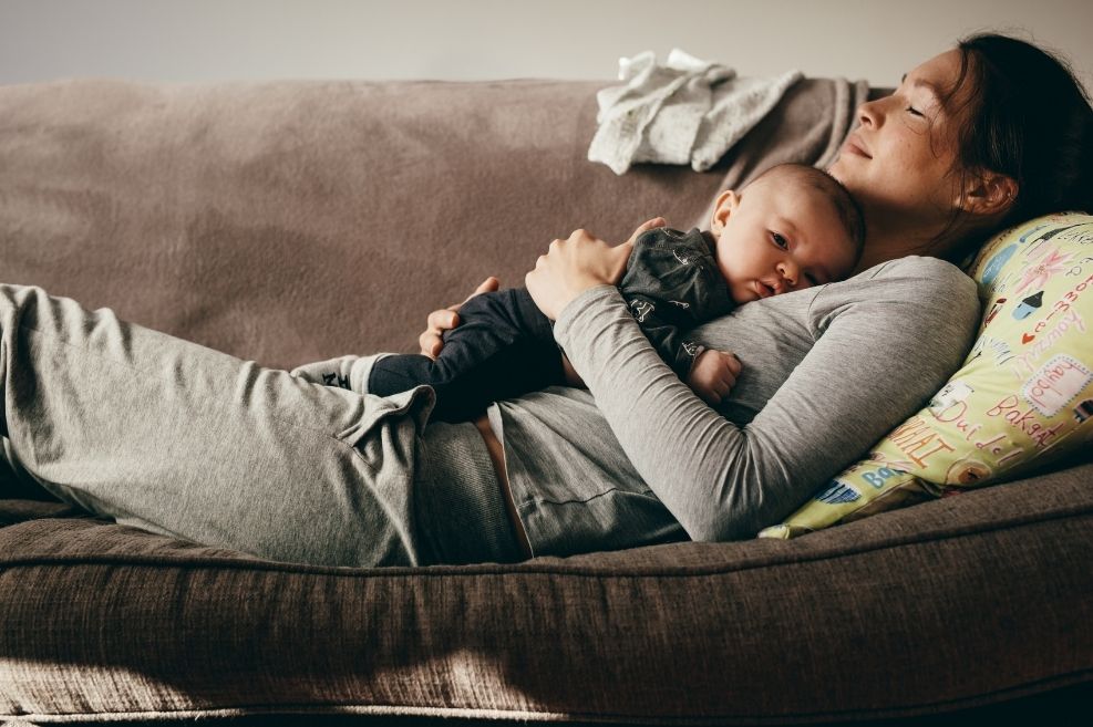 A woman and a baby peacefully resting on a brown couch, after enjoying the benefits of a postpartum care package.