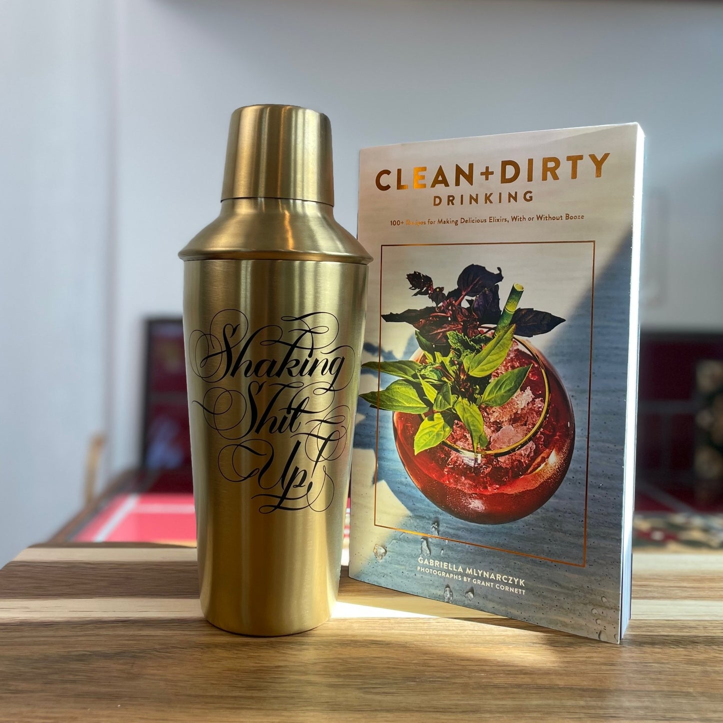 The Clean + Dirty Drinking recipe book next to the gold cocktail shaker on a kitchen counter